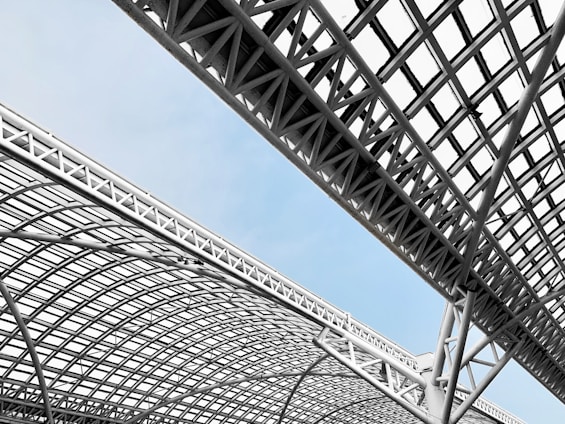 A complex and modern architectural structure with crisscrossing metal beams and latticework against a light blue sky. The design features geometric shapes and angles, creating a dynamic and industrial aesthetic.