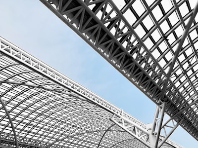 A complex and modern architectural structure with crisscrossing metal beams and latticework against a light blue sky. The design features geometric shapes and angles, creating a dynamic and industrial aesthetic.
