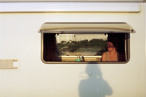 A woman is sitting inside a camper van, visible through an open window. The interior appears cozy with various items on the windowsill, and there is a shadow of a person holding a camera on the exterior wall of the van. The background outside the window shows some greenery.