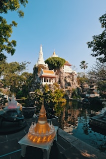 A tranquil scene featuring a garden with lush greenery surrounding a small, serene pond. In the distance, there&rsquo;s a stunning temple structure intricately built atop a rocky formation, partially covered with green foliage. In the foreground, a decorative illuminated model of a pagoda sits on a table, adding a warm glow to the setting. The sky above is clear and blue, contributing to the peaceful ambiance.