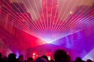 An architectural structure is illuminated with vibrant laser beams in shades of red and purple. The lasers converge at a central point, creating a captivating display that frames the building's intricate facade. Silhouettes of people are visible in the foreground, observing the light show.
