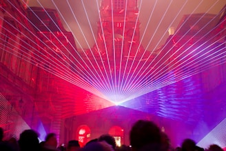 An architectural structure is illuminated with vibrant laser beams in shades of red and purple. The lasers converge at a central point, creating a captivating display that frames the building's intricate facade. Silhouettes of people are visible in the foreground, observing the light show.