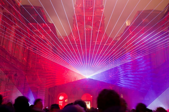 An architectural structure is illuminated with vibrant laser beams in shades of red and purple. The lasers converge at a central point, creating a captivating display that frames the building's intricate facade. Silhouettes of people are visible in the foreground, observing the light show.