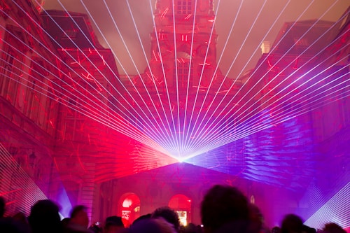 An architectural structure is illuminated with vibrant laser beams in shades of red and purple. The lasers converge at a central point, creating a captivating display that frames the building's intricate facade. Silhouettes of people are visible in the foreground, observing the light show.
