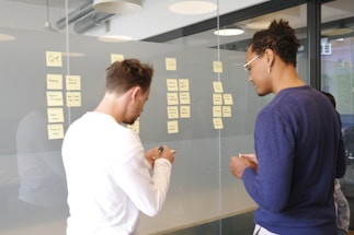 Two individuals are seen in a modern office setting, engaging in a task that involves writing and arranging sticky notes on a glass wall. The sticky notes have various handwritten notes on them, possibly related to project planning or brainstorming. The room has large windows and modern lighting fixtures.