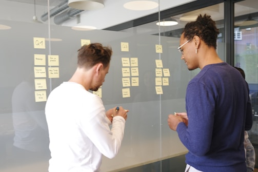 Two individuals are seen in a modern office setting, engaging in a task that involves writing and arranging sticky notes on a glass wall. The sticky notes have various handwritten notes on them, possibly related to project planning or brainstorming. The room has large windows and modern lighting fixtures.