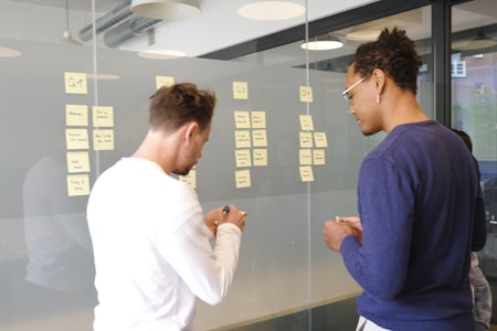 Two individuals are seen in a modern office setting, engaging in a task that involves writing and arranging sticky notes on a glass wall. The sticky notes have various handwritten notes on them, possibly related to project planning or brainstorming. The room has large windows and modern lighting fixtures.