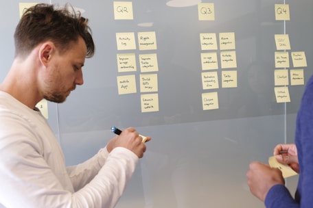 Several people are collaborating in an office setting, using sticky notes on a transparent board divided into columns labeled Q2, Q3, and Q4. Each column contains tasks or goals labeled under various headings. One person is writing on a sticky note while another is holding notes.