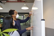 Two people wearing masks work collaboratively on a large whiteboard in an office setting. They are focused on writing and discussing ideas, with one person holding a marker and actively writing while the other observes closely. Overhead lights illuminate the workspace, and there is a plant in the foreground.