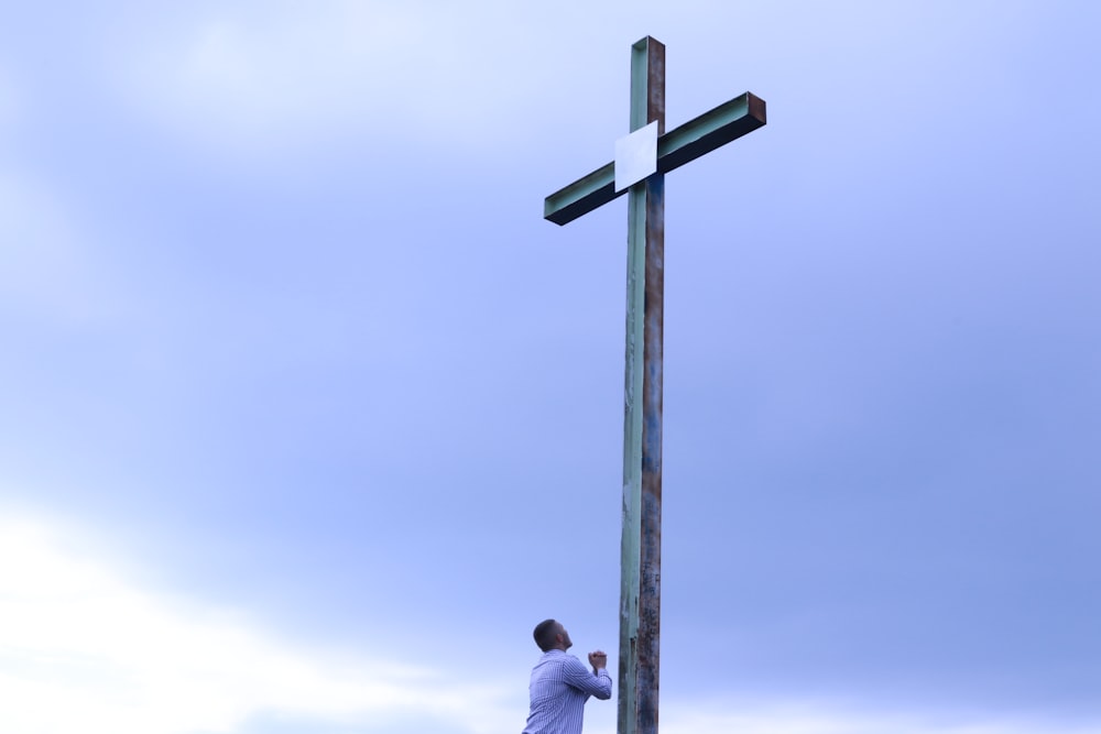 homme en chemise blanche debout à côté d’une croix en bois marron sous des nuages blancs pendant la journée