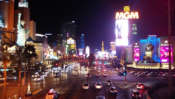 A bustling nightlife scene in an urban area with bright neon lights, high-rise buildings, and heavy traffic. The image includes illuminated signs, palm trees, and replicas of iconic landmarks.