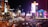 A bustling nightlife scene in an urban area with bright neon lights, high-rise buildings, and heavy traffic. The image includes illuminated signs, palm trees, and replicas of iconic landmarks.