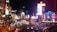 A bustling nightlife scene in an urban area with bright neon lights, high-rise buildings, and heavy traffic. The image includes illuminated signs, palm trees, and replicas of iconic landmarks.