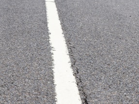 A close-up of fresh asphalt being laid on a sunny day with construction cones nearby.