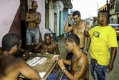 Several men are gathered around a small table playing dominoes on a street. Most of them are shirtless, and one of them is wearing a yellow shirt. The atmosphere has a casual and social vibe with people engaging in conversation and leisure activities. There is a bicycle leaning against the wall, and the setting appears to be in a residential area with a few buildings in the background.