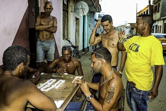 Several men are gathered around a small table playing dominoes on a street. Most of them are shirtless, and one of them is wearing a yellow shirt. The atmosphere has a casual and social vibe with people engaging in conversation and leisure activities. There is a bicycle leaning against the wall, and the setting appears to be in a residential area with a few buildings in the background.