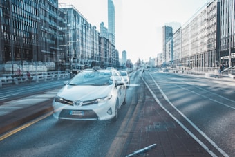 A city street scene with a white taxi in the foreground. The street is surrounded by tall, modern buildings with glass facades on both sides. The long exposure effect creates a blurred, dreamy look with overlapping images. The taxi is positioned prominently in the center with some motion trails. The scene is mostly empty with a few distant pedestrians.