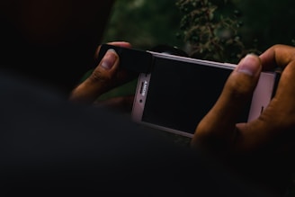 A person is holding a smartphone and what appears to be an attachment for photography, possibly a lens, is being fitted over the phone's camera. The smartphone screen is dark, and the background includes blurry vegetation, creating a moody and focused setting.