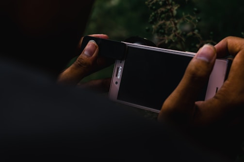 A person is holding a smartphone and what appears to be an attachment for photography, possibly a lens, is being fitted over the phone's camera. The smartphone screen is dark, and the background includes blurry vegetation, creating a moody and focused setting.