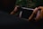 A person is holding a smartphone and what appears to be an attachment for photography, possibly a lens, is being fitted over the phone's camera. The smartphone screen is dark, and the background includes blurry vegetation, creating a moody and focused setting.