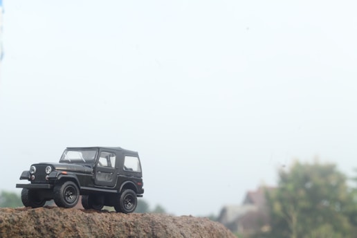 A small model of a black jeep is placed on a rough surface, resembling rocky terrain. The background is blurred with hints of greenery and buildings, creating an outdoor or adventure setting.