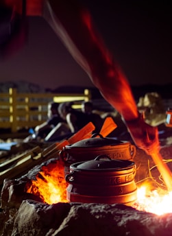 A cooking scene in an outdoor setting at night where cast iron pots are placed over a fire pit. The flames cast a warm glow on the pots and the surrounding area. In the background, there are silhouettes of people seated, suggesting a group gathering or camping atmosphere.