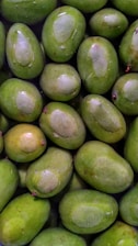 A collection of fresh, green mangoes closely packed together. The mangoes have smooth skin with patches of lighter green and are arranged in an overlapping pattern. Some mangoes have small, protruding stems visible.
