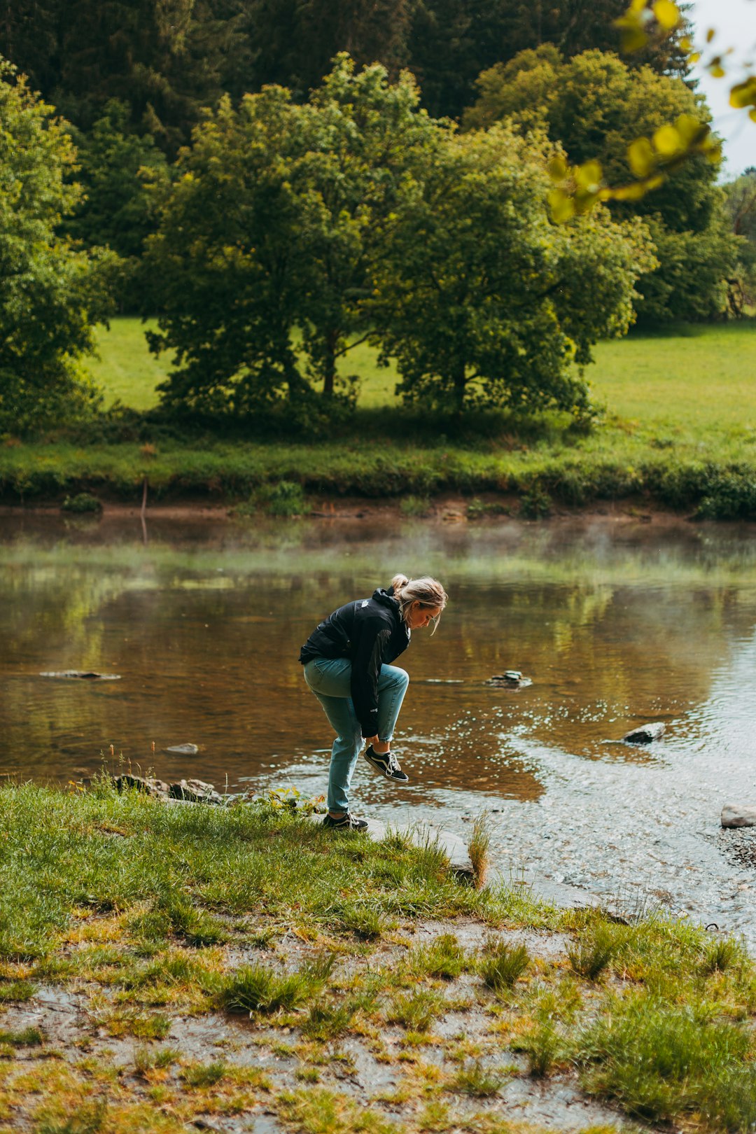 man in black jacket and blue denim jeans standing on green grass field near river during