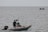 A person stands on a small inflatable boat with a motor, positioned on a vast body of water. The boat appears to have a small flag, and in the background, another boat with people is visible far on the horizon.