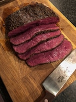 Several slices of cooked beef roast are laid out on a wooden cutting board. The meat is medium rare, with a pink interior and a browned crust. A large knife with a wooden handle, featuring some moisture, rests beside the sliced meat on the board.