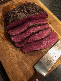 Several slices of cooked beef roast are laid out on a wooden cutting board. The meat is medium rare, with a pink interior and a browned crust. A large knife with a wooden handle, featuring some moisture, rests beside the sliced meat on the board.