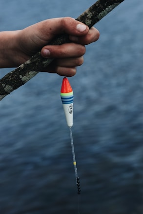 A hand holding a fishing rod, with a colorful fishing float attached, is positioned over a body of water. The float features red, blue, green, and yellow bands. The water in the background appears calm, suggesting a serene fishing setting.