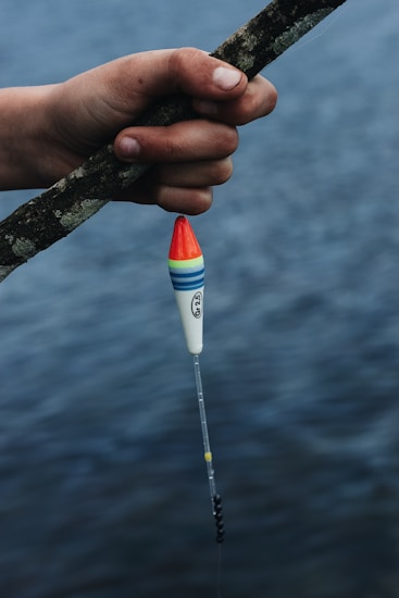 A hand holding a fishing rod, with a colorful fishing float attached, is positioned over a body of water. The float features red, blue, green, and yellow bands. The water in the background appears calm, suggesting a serene fishing setting.
