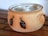 A ceramic pot with two handles and a lid, decorated with painted images of black birds. The pot is placed on a wooden surface and has a natural earthy color.