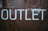 Large white letters spell out the word 'OUTLET' against a dark background with a mesh or chain-link fence pattern.