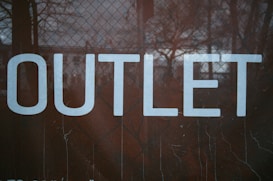 Large white letters spell out the word 'OUTLET' against a dark background with a mesh or chain-link fence pattern.