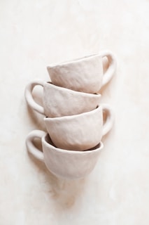 Four rustic, unglazed clay cups are stacked on top of each other against a gentle, creamy background. The texture of the cups appears rough, highlighting their handcrafted nature.