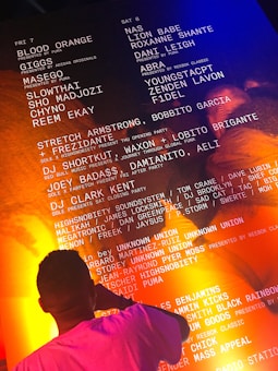 A person in a pink shirt is standing in front of a large illuminated poster featuring a list of performers and events, possibly for a music festival or concert series. The background has a combination of orange and blue lighting. The text includes names of artists such as Blood Orange, Nas, and others, along with sponsors like Adidas and Puma.