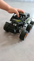 A child’s hand is placed on top of a black remote-controlled toy car with large, rugged tires and green and white decals. The car has a robust, off-road design, and is positioned on a smooth, light-colored surface.