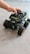A child’s hand is placed on top of a black remote-controlled toy car with large, rugged tires and green and white decals. The car has a robust, off-road design, and is positioned on a smooth, light-colored surface.