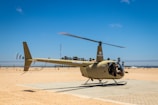 A helicopter is parked on a flat, sandy surface under a clear blue sky. It is beige in color and features the markings 'V5-HRA' and 'R44' on its tail. The area is open and expansive, with a fence and scattered palm trees in the background. The scene conveys a sense of quietness and isolation, with only the helicopter as the main subject.