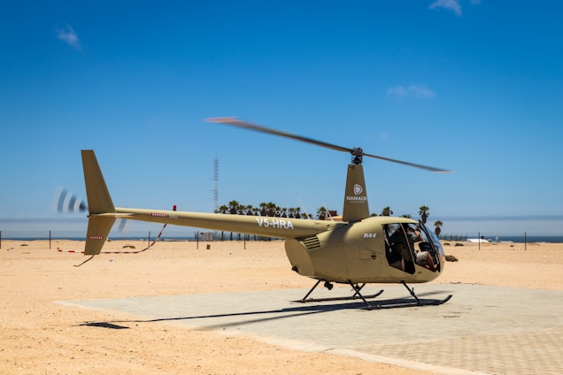 A helicopter is parked on a flat, sandy surface under a clear blue sky. It is beige in color and features the markings 'V5-HRA' and 'R44' on its tail. The area is open and expansive, with a fence and scattered palm trees in the background. The scene conveys a sense of quietness and isolation, with only the helicopter as the main subject.