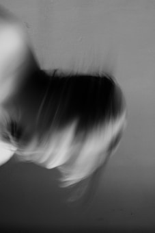 A blurred black and white image showing motion, possibly of a person's head or hair moving rapidly against a plain background.