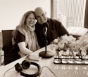 A smiling couple is seated closely together, with arms around each other, in a modern office or studio setting. A table with a microphone, headphones, and a smartphone are in front of them. A window with a city view is in the background, and there are vases with flowers on the table.