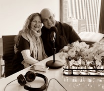 A smiling couple is seated closely together, with arms around each other, in a modern office or studio setting. A table with a microphone, headphones, and a smartphone are in front of them. A window with a city view is in the background, and there are vases with flowers on the table.