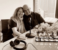 A smiling couple is seated closely together, with arms around each other, in a modern office or studio setting. A table with a microphone, headphones, and a smartphone are in front of them. A window with a city view is in the background, and there are vases with flowers on the table.