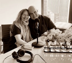 A smiling couple is seated closely together, with arms around each other, in a modern office or studio setting. A table with a microphone, headphones, and a smartphone are in front of them. A window with a city view is in the background, and there are vases with flowers on the table.