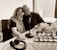 A smiling couple is seated closely together, with arms around each other, in a modern office or studio setting. A table with a microphone, headphones, and a smartphone are in front of them. A window with a city view is in the background, and there are vases with flowers on the table.