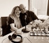 A smiling couple is seated closely together, with arms around each other, in a modern office or studio setting. A table with a microphone, headphones, and a smartphone are in front of them. A window with a city view is in the background, and there are vases with flowers on the table.