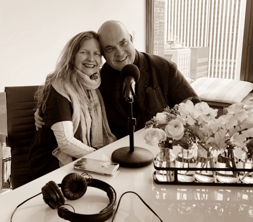 A smiling couple is seated closely together, with arms around each other, in a modern office or studio setting. A table with a microphone, headphones, and a smartphone are in front of them. A window with a city view is in the background, and there are vases with flowers on the table.